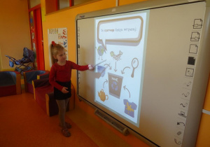 Dziewczynka stoi pod tablicą interaktywną i wskaźnikiem pokazuje szkielet ryby.